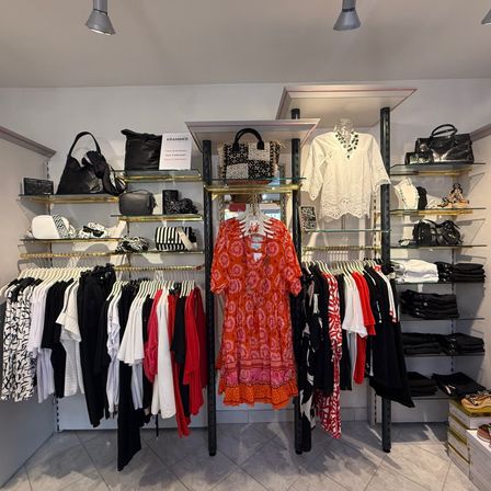 Ein Bekleidungsgeschäft mit ordentlich angeordneten Kleidern an Kleiderbügeln und Regalen. Taschen, eine weiße Bluse und ein rotes Kleid sind ausgestellt. Der Laden hat weiße Wände und Decke mit Lampen.