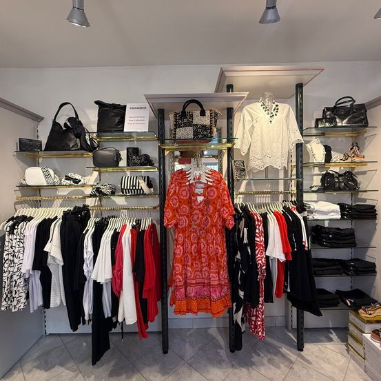 Ein Bekleidungsgeschäft mit ordentlich angeordneten Kleidern an Kleiderbügeln und Regalen. Taschen, eine weiße Bluse und ein rotes Kleid sind ausgestellt. Der Laden hat weiße Wände und Decke mit Lampen.