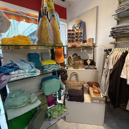 Ein Bekleidungsgeschäft zeigt verschiedene Taschen und Accessoires auf Glasregalen. Eine Wand mit gefalteten Kleidern und eine offene Tür führen nach draußen.