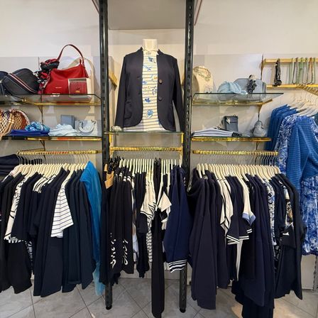 Ein Bekleidungsgeschäft mit einem weißen und blauen Farbschema, das eine Vielzahl von Outfits präsentiert. Mannequins zeigen Jacken und Hemden. Regale halten Handtaschen, Schuhe und Accessoires. Der Boden ist gefliest.