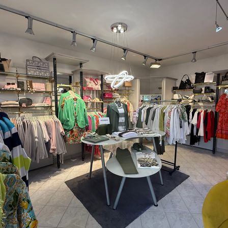 Ein Boutique zeigt eine Vielzahl von Kleidungsstücken. Eine kreisförmige Lampe hängt von der Decke, und Kleidung hängt an Kleiderbügeln. Ein Tisch in der Mitte hat Gegenstände darauf. Regale mit Taschen sind an der Wand.