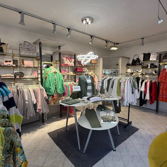 Ein Boutique zeigt eine Vielzahl von Kleidungsstücken. Eine kreisförmige Lampe hängt von der Decke, und Kleidung hängt an Kleiderbügeln. Ein Tisch in der Mitte hat Gegenstände darauf. Regale mit Taschen sind an der Wand.