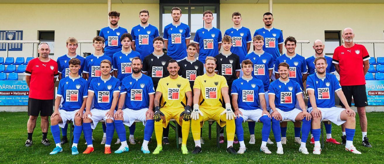Die Fußballmannschaft von SV WeiKersdorf steht vor ihrem Stadion. Alle Spieler tragen ihre Teamtrikots und posieren für ein Mannschaftsfoto. Das Stadion hat ein Schild, das 'SV WeiKersdorf' steht.