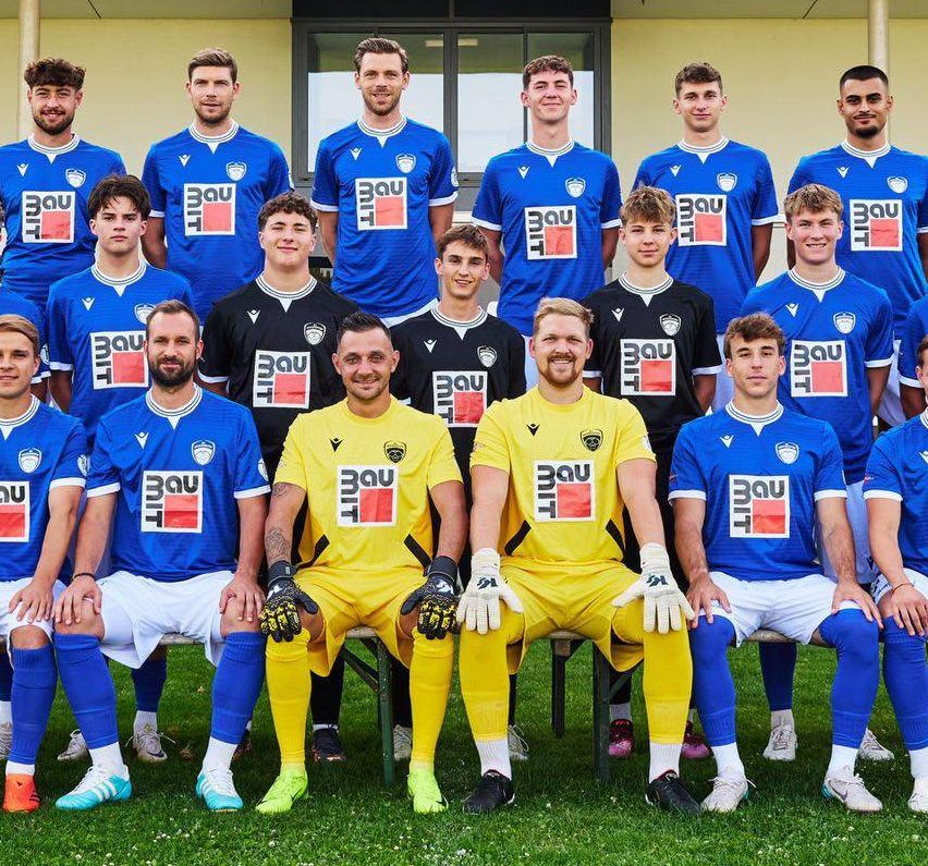 Die Fußballmannschaft von SV WeiKersdorf steht vor ihrem Stadion. Alle Spieler tragen ihre Teamtrikots und posieren für ein Mannschaftsfoto. Das Stadion hat ein Schild, das 'SV WeiKersdorf' steht.