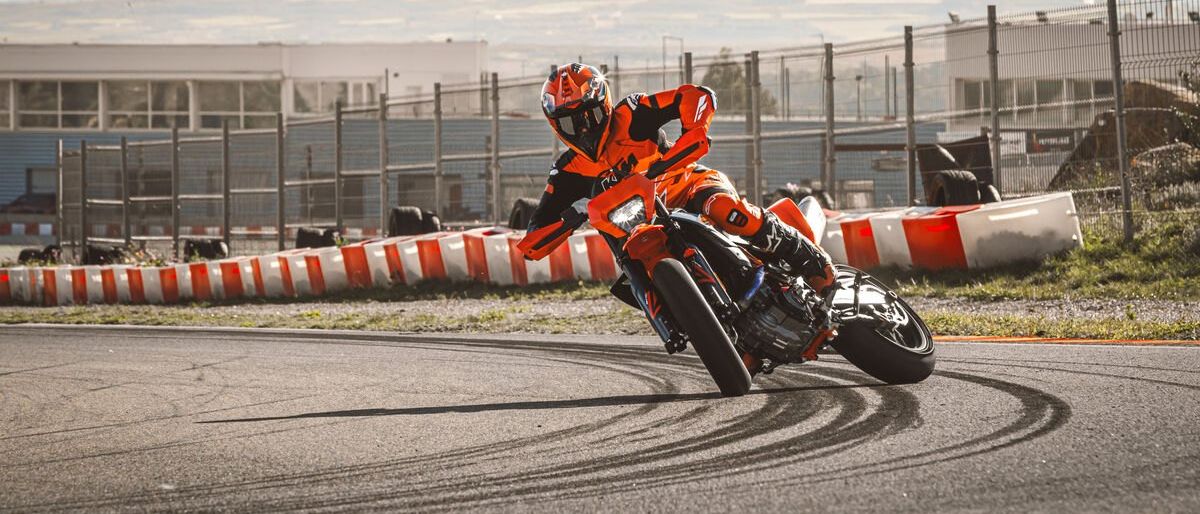 Ein Motorradfahrer in orangefarbener Kleidung fährt auf einer Rennstrecke. Der Fahrer lehnt sich in eine Kurve mit Bergen im Hintergrund.