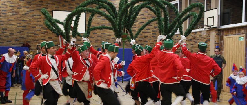 A group of men in red uniforms and green hats dance in a circle around a large green Christmas tree in a hall.