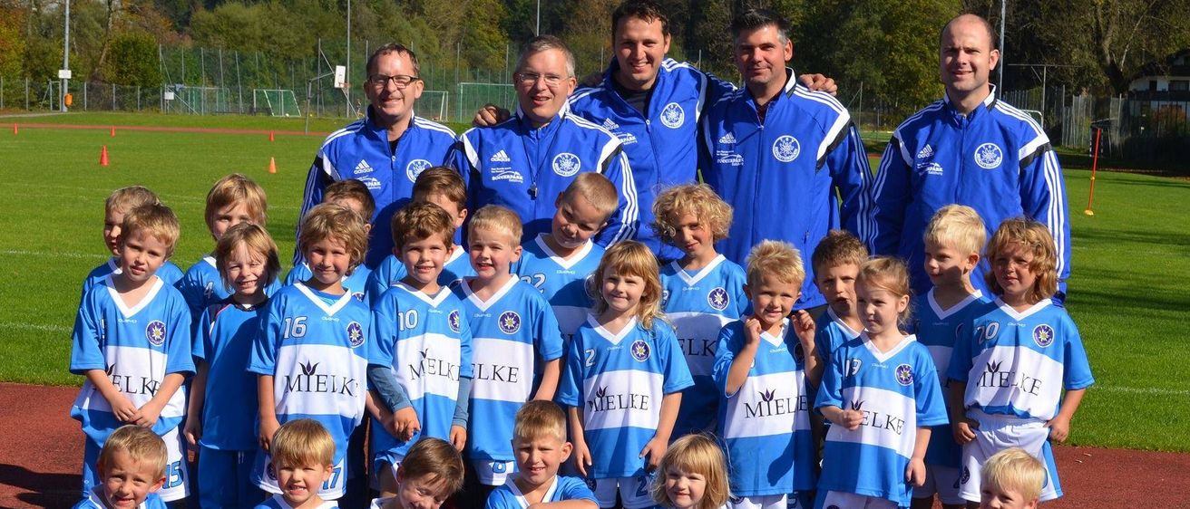 Eine Jugendfußballmannschaft in blauen Trikots mit weißen Sternen posiert für ein Foto auf einem Feld mit Bäumen und Bergen im Hintergrund.
