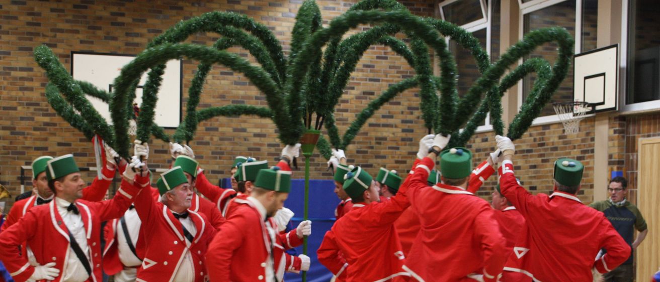 Eine Gruppe von Männern in roten Uniformen mit grünen Hüten tanzt um eine große grüne Struktur herum, möglicherweise während einer festlichen Veranstaltung in einer Halle.