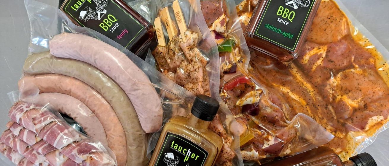 Verschiedene Packungen mit Fleisch und Würsten, begleitet von mehreren Flaschen mit Etiketten wie 'Taucher BBQ' und 'Taucher Mangofruchtig', sind auf einem Tablett angeordnet. Das Fleisch und die Würste befinden sich in durchsichtigen Plastiktüten.