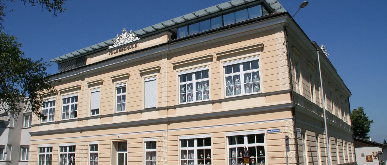 Ein großes Gebäude mit mehreren Fenstern, einer weißen Tür und einer Dachterrasse. Das Gebäude trägt ein Schild mit der Aufschrift 'Volksschule'.