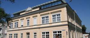 Ein großes Gebäude mit mehreren Fenstern, einer weißen Tür und einer Dachterrasse. Das Gebäude trägt ein Schild mit der Aufschrift 'Volksschule'.