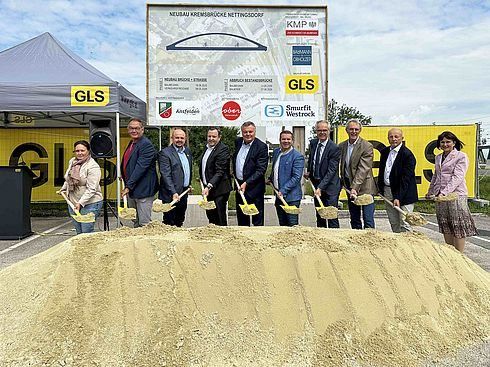 Eine Gruppe von Menschen in formeller Kleidung steht um einen Haufen Erde herum und hält Schaufeln. Sie befinden sich auf einer Baustelle mit einem gelben Banner.