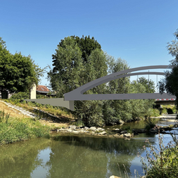 Eine graue Bogenbrücke überspannt einen Fluss, umgeben von üppigem Grün und Bäumen auf beiden Seiten unter einem klaren blauen Himmel.