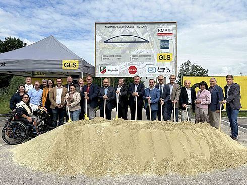 Eine Gruppe von Menschen steht um einen Haufen Erde herum und hält Schaufeln, wahrscheinlich für eine Grundsteinlegungszeremonie. Sie stehen vor einem großen Schild mit Text und Logos. Ein Mensch im Rollstuhl befindet sich auf der linken Seite. Ein Zeltdach befindet sich auf der linken Seite.