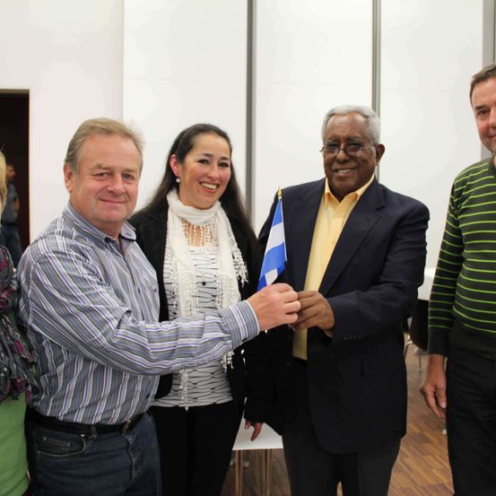 Vier Menschen stehen lächelnd für ein Foto. Ein Mann links hält eine Flagge und ein anderer Mann rechts trägt ein gestreiftes Hemd.