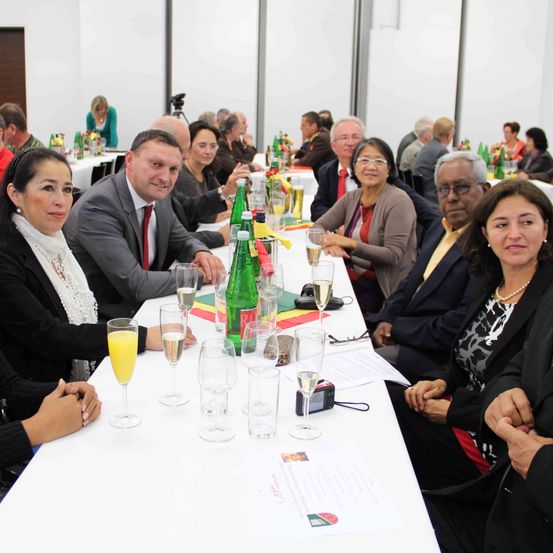 Eine Gruppe von Menschen, wahrscheinlich Kollegen, sitzt an einem langen Tisch mit verschiedenen Getränken und Flaschen. Einige halten Weingläser, während andere Gläser mit Saft haben. Eine Frau in der Vorderfront lächelt, während ihre Hände auf dem Tisch ruhen.