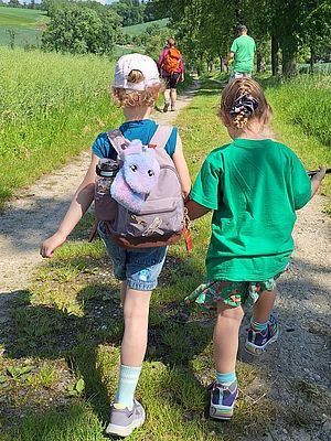 Zwei Kinder gehen gemeinsam auf einem Pfad durch ein Grasfeld. Ein Rucksack mit einem Kuscheltier befindet sich auf dem Rücken des Kindes auf der linken Seite.