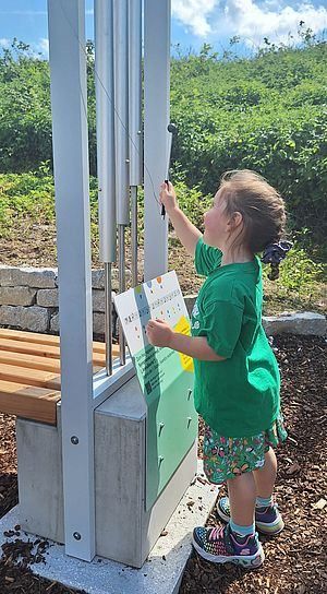 Ein kleines Kind in einem grünen Shirt spielt mit einer Windglocke in einem Park. Die Windglocke ist an einem grünen und grauen Pfosten befestigt.