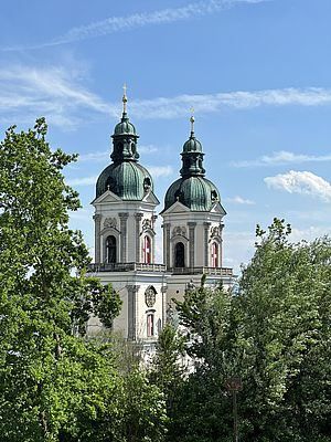 Zwei hohe Türme mit grünen Kuppeln sind von Bäumen umgeben. Die Türme haben ein weißes und beiges Design mit roten Akzenten. Der Himmel darüber ist blau mit einigen Wolken.