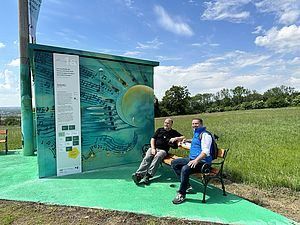 Zwei Männer sitzen auf einer Bank vor einer bemalten Struktur. Sie sind lässig gekleidet und scheinen für ein Foto zu posieren. Dahinter befindet sich ein grasbewachsenes Feld mit Bäumen.
