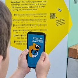 Eine Person hält ein Handy vor einem Poster, das Noten und Text hat. Das Poster hat ein Bild eines QR-Codes, und das Handy hat eine offene App. Das Poster hat einen gelben Hintergrund.