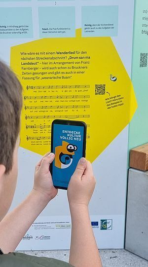 Eine Person hält ein Handy vor einem Poster, das Noten und Text hat. Das Poster hat ein Bild eines QR-Codes, und das Handy hat eine offene App. Das Poster hat einen gelben Hintergrund.