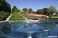 Ein ruhiger Wasserbereich, umgeben von Grün, mit einer roten Rutsche auf einem Hügel im Hintergrund.