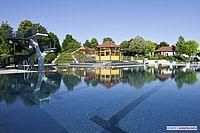 Ein großer Swimmingpool mit Sprungbrett und Wasserrutsche spiegelt die Umgebung wider, einschließlich eines Pavillons, Häusern und Bäumen.