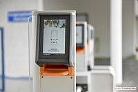 Mehrere Selbstbedienungs-Kioske sind in einem Flur aufgereiht, jeder mit einem Bildschirm, der eine digitale Benutzeroberfläche anzeigt. Die Bildschirme sind beleuchtet und zeigen eine Benutzeroberfläche mit Symbolen und Text.