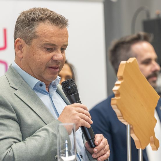 Ein Mann in einem grauen Anzug hält ein Mikrofon und spricht bei einer Veranstaltung. Hinter ihm lauscht ein anderer Mann. In der Nähe steht ein hölzernes Ausschnitt einer Karte.