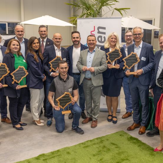 Gruppenfoto von Personen, die Auszeichnungen in einem Konferenzsaal halten. Ein Mann in Anzug kniet vorne und hält seine Auszeichnung. Hinter ihnen ist ein Banner zu sehen.