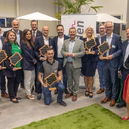 Gruppenfoto von Personen, die Auszeichnungen in einem Konferenzsaal halten. Ein Mann in Anzug kniet vorne und hält seine Auszeichnung. Hinter ihnen ist ein Banner zu sehen.