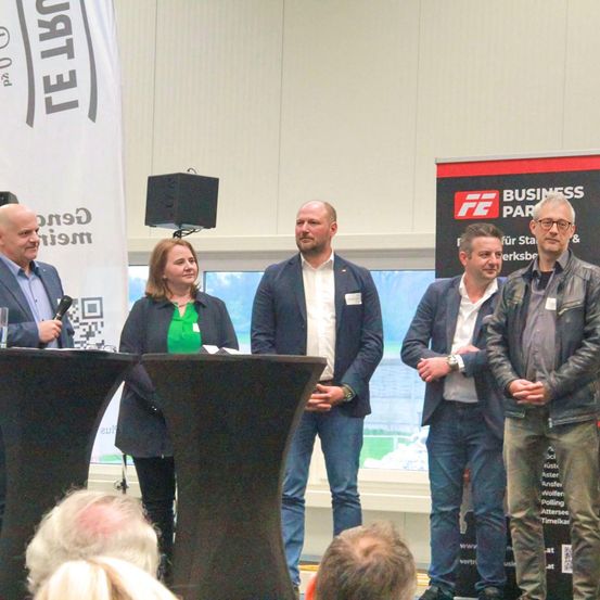 Fünf Personen stehen bei einer Veranstaltung zusammen. Die Frau links hält ein Mikrofon. Hinter ihnen befindet sich ein großes Banner mit der Aufschrift "Business Park".