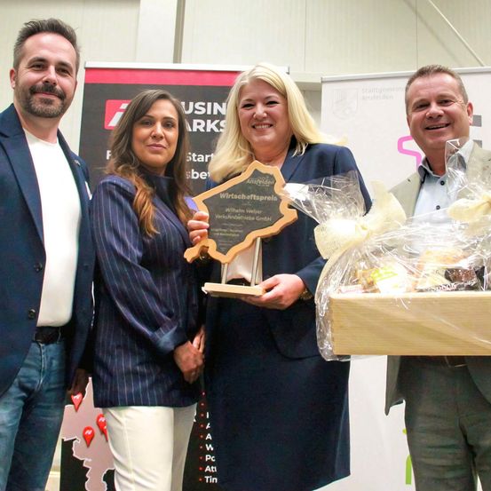 Vier Menschen stehen lächelnd zusammen. Zwei Frauen halten einen Preis und einen Korb. Die Frauen tragen Anzüge. Die Männer tragen Anzüge und Krawatten. Die Umgebung erscheint professionell.