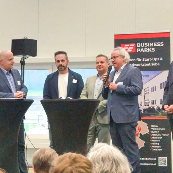 Fünf Männer in Anzügen stehen an einem Rednerpult mit einem Mikrofon. Ein Mann spricht. Ein Banner im Hintergrund sagt 'BUSINESS PARKS'.