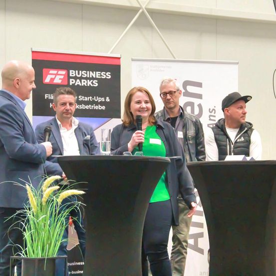 Fünf Personen stehen vor einem Banner auf einem Podium und lächeln. Eine Frau hält ein Mikrofon und ein Mann neben ihr einen Glas. Ein Mann zu ihrer Linken hält ein Mikrofon. Eine Pflanze ist im Vordergrund.