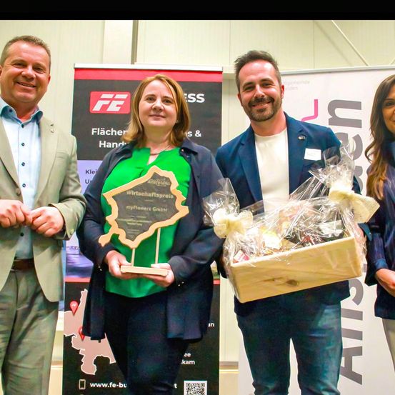 Eine Gruppe von vier Personen steht zusammen. Eine Frau hält eine Trophäe und ein Mann einen Geschenkkorb. Sie alle lächeln.