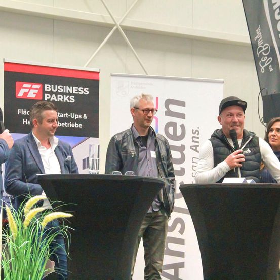 Drei Männer in Business-Kleidung stehen an einem Podium mit einer Frau. Sie lächeln und blicken in die Kamera. Hinter ihnen befinden sich zwei Banner.