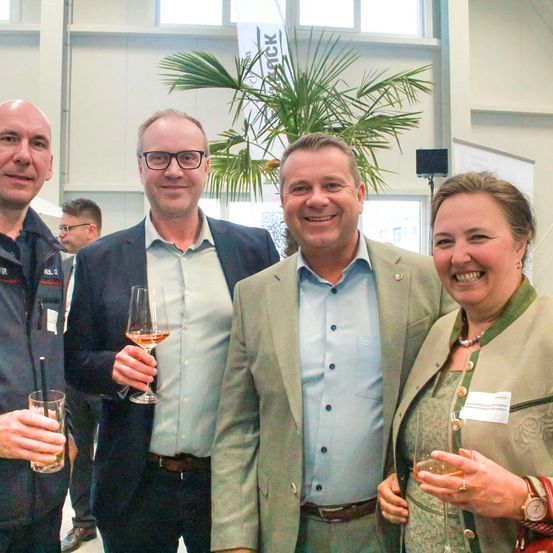 Vier Menschen lächeln und posieren für ein Foto bei einer Veranstaltung. Ein Mann hält ein Glas Wein. Hinter ihnen steht eine Pflanze und ein Banner.