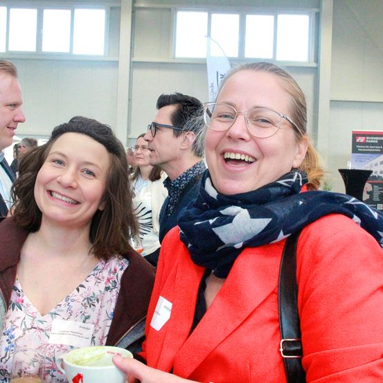 Zwei Frauen lächeln und lachen gemeinsam bei einer Geschäftsveranstaltung. Die Frau auf der rechten Seite trägt eine Brille und einen roten Blazer. Im Hintergrund unterhalten sich mehrere Personen.