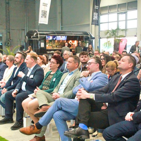Eine Gruppe von Fachleuten in formeller Kleidung sitzt aufmerksam auf Stühlen in einem großen Saal. Sie konzentrieren sich auf die Vorderseite, möglicherweise auf eine Präsentation. Der Raum hat Glasfenster und einen geparkten Imbisswagen im Hintergrund.