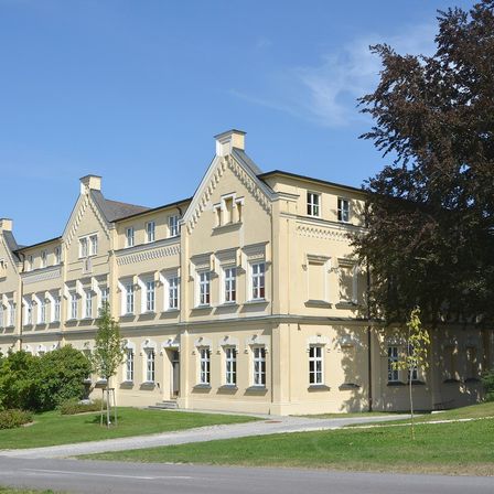 Ein großes beiges Gebäude mit mehreren Fenstern steht auf einem Rasen unter einem klaren blauen Himmel, umgeben von Bäumen und Sträuchern.