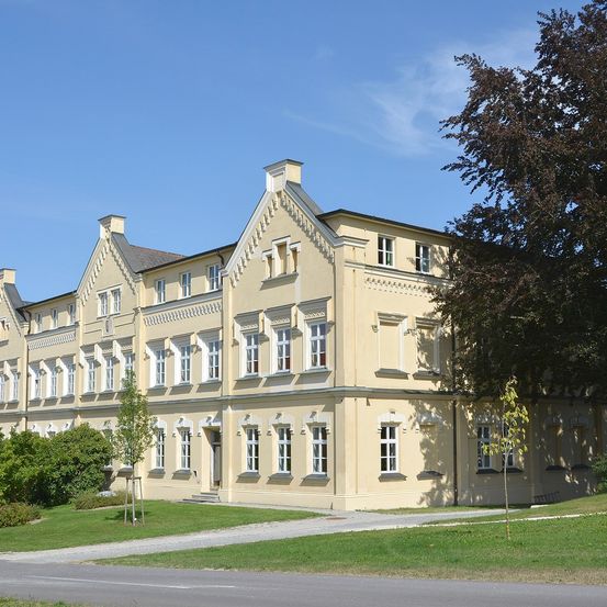 Ein großes beiges Gebäude mit mehreren Fenstern steht auf einem Rasen unter einem klaren blauen Himmel, umgeben von Bäumen und Sträuchern.