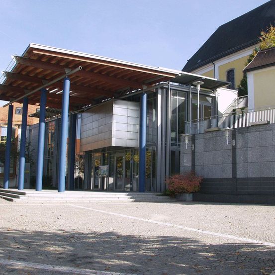 Ein modernes Gebäude mit einem Holzdach und blauen Säulen, Glastüren und Fenstern. Ein Topf mit Pflanzen steht vor dem Gebäude.