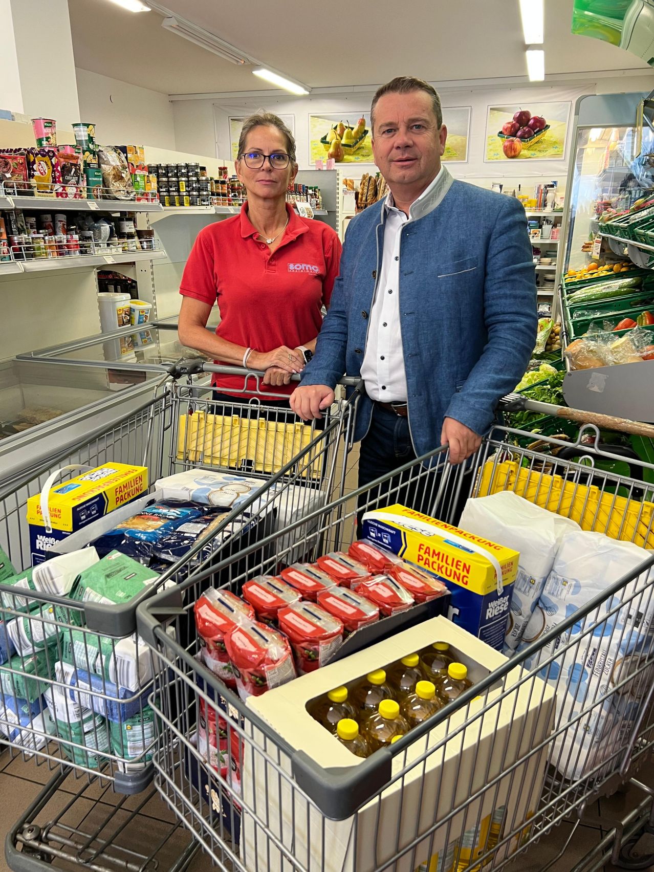 Ein Mann und eine Frau stehen hinter einem mit Lebensmitteln gefüllten Einkaufswagen in einem Geschäft. Die Frau trägt ein rotes Shirt mit dem Wort 'SOMA' darauf.
