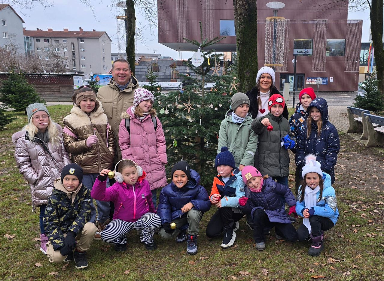 Eine Gruppe von Kindern und Erwachsenen, die Winterkleidung tragen, posiert für ein Foto vor zwei Weihnachtsbäumen. Sie befinden sich in einem Park mit einem Gebäude im Hintergrund.