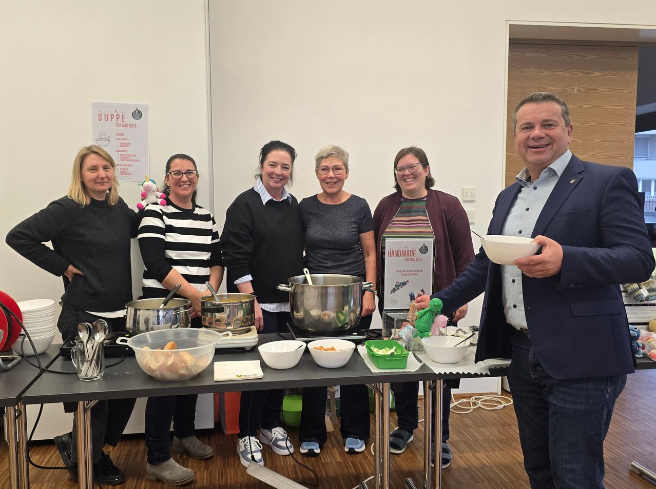 Six women and a man are smiling and posing for a photo. They are standing in front of a table filled with food in bowls and pots. Behind them is a sign that reads 'Suppe'.