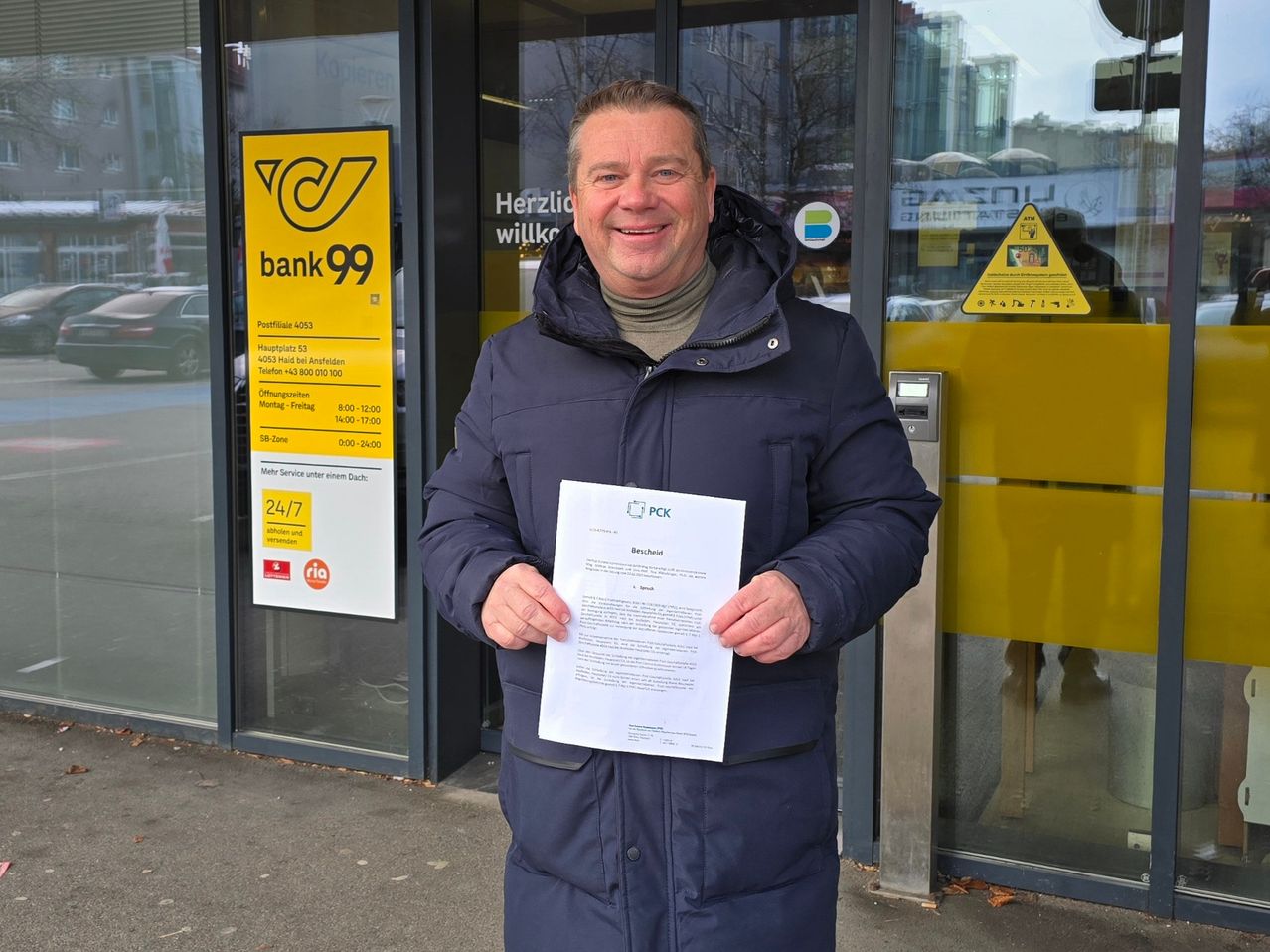 Ein Mann steht lächelnd vor dem Eingang einer Bank und hält ein Stück Papier in der Hand. Er trägt einen blauen Mantel. Hinter ihm zeigt ein Schild die Öffnungszeiten der Bank an.