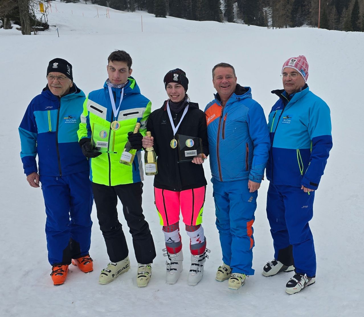 Fünf Erwachsene stehen im Schnee. Sie tragen Winterkleidung und halten Champagnerflaschen. Sie lächeln und scheinen für ein Foto zu posieren.