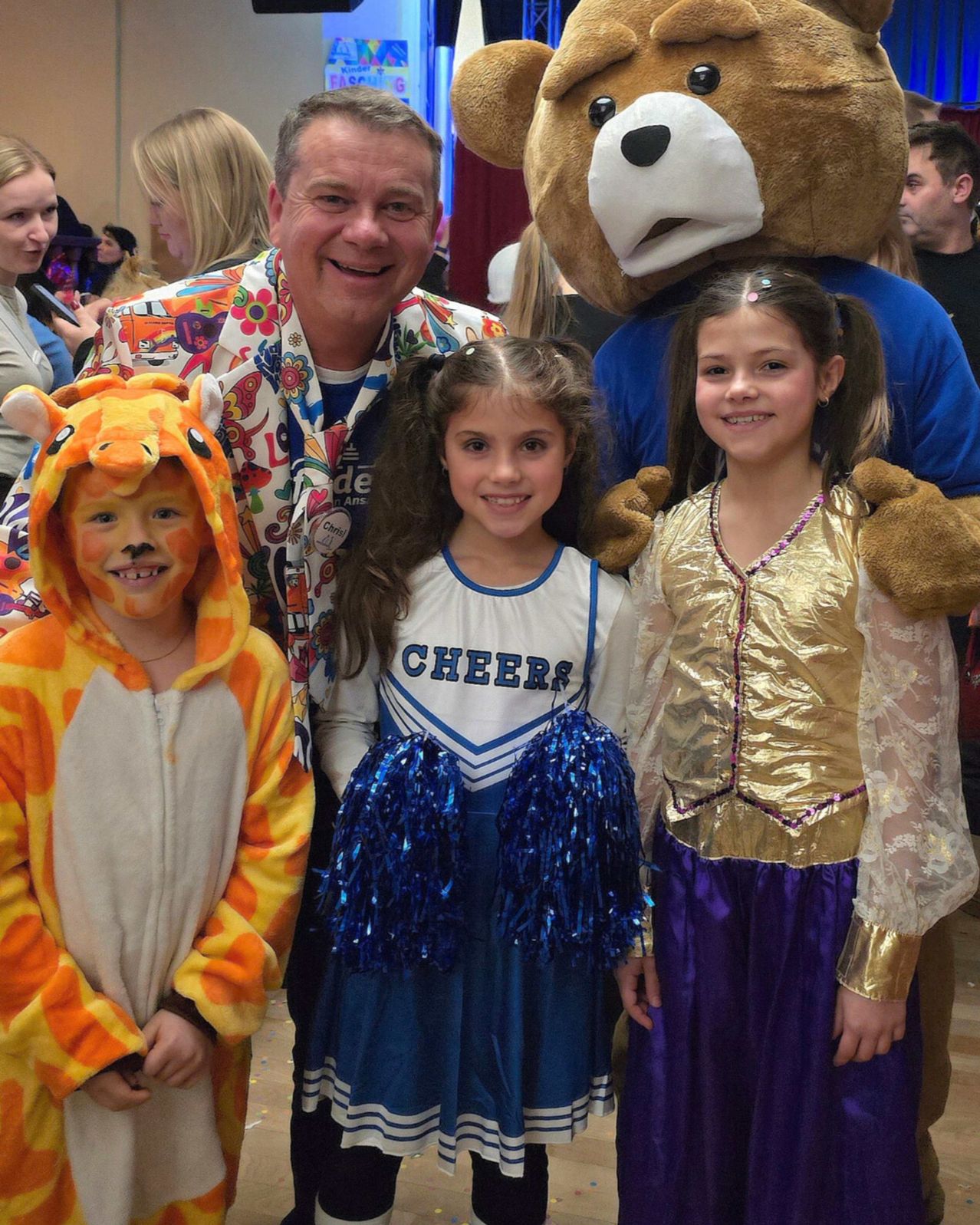 Ein Mann und zwei Mädchen in Kostümen posieren mit einem Bärenmaskottchen. Der Mann trägt ein Blumenhemd. Die Mädchen tragen Cheerleader- und Prinzessinnenkostüme. Der Hintergrund ist unscharf.